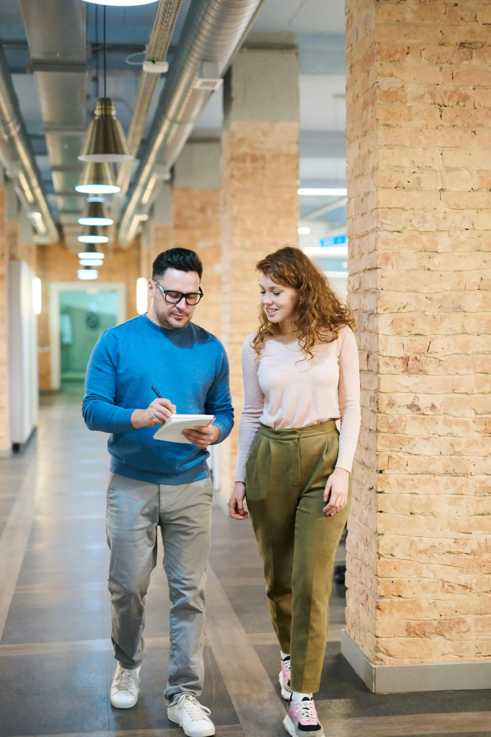 Two colleagues walking through a bright hallway while discussing notes